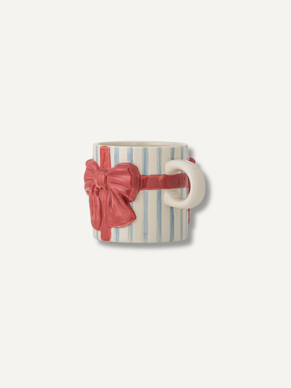 A ceramic mug featuring a white and light blue vertical stripe pattern, wrapped in a 3D red ribbon with a large red bow, set against a plain off-white background.