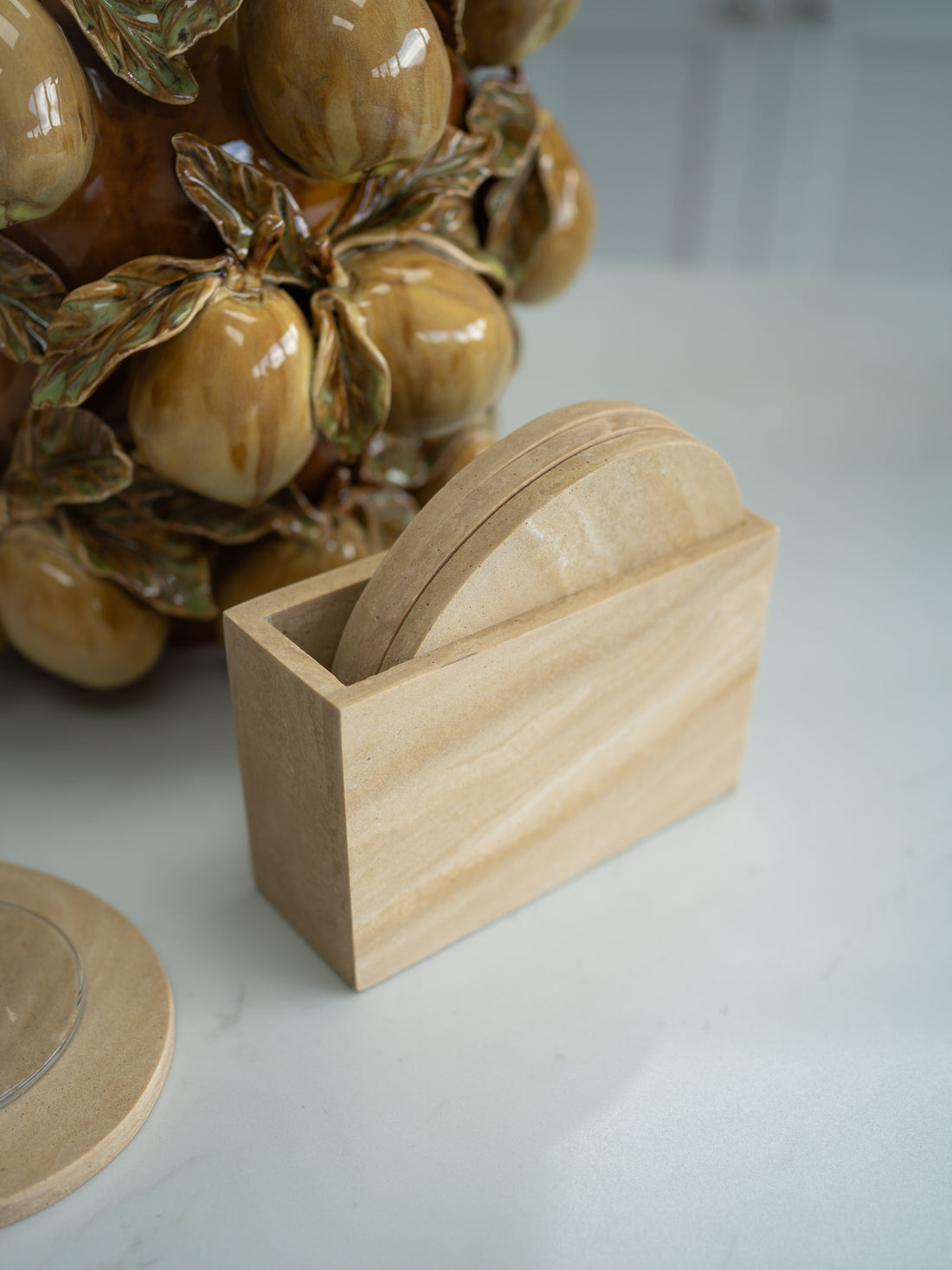 Travertine effect coasters on a marble surface with a ceramic fruit sculpture in the background.