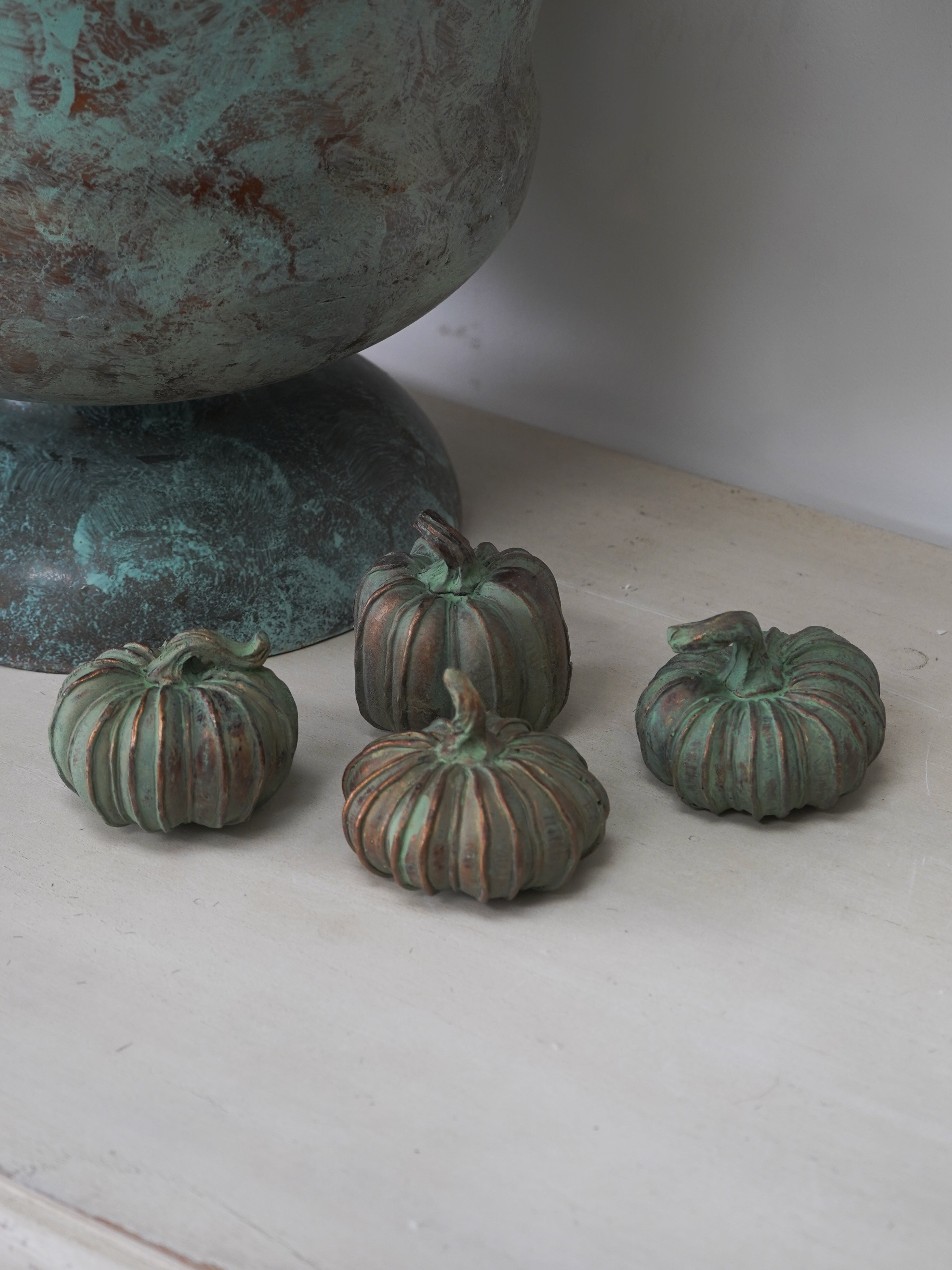 Four small green copper-colored decorative pumpkins on a white surface with a large textured bronze object in the background.