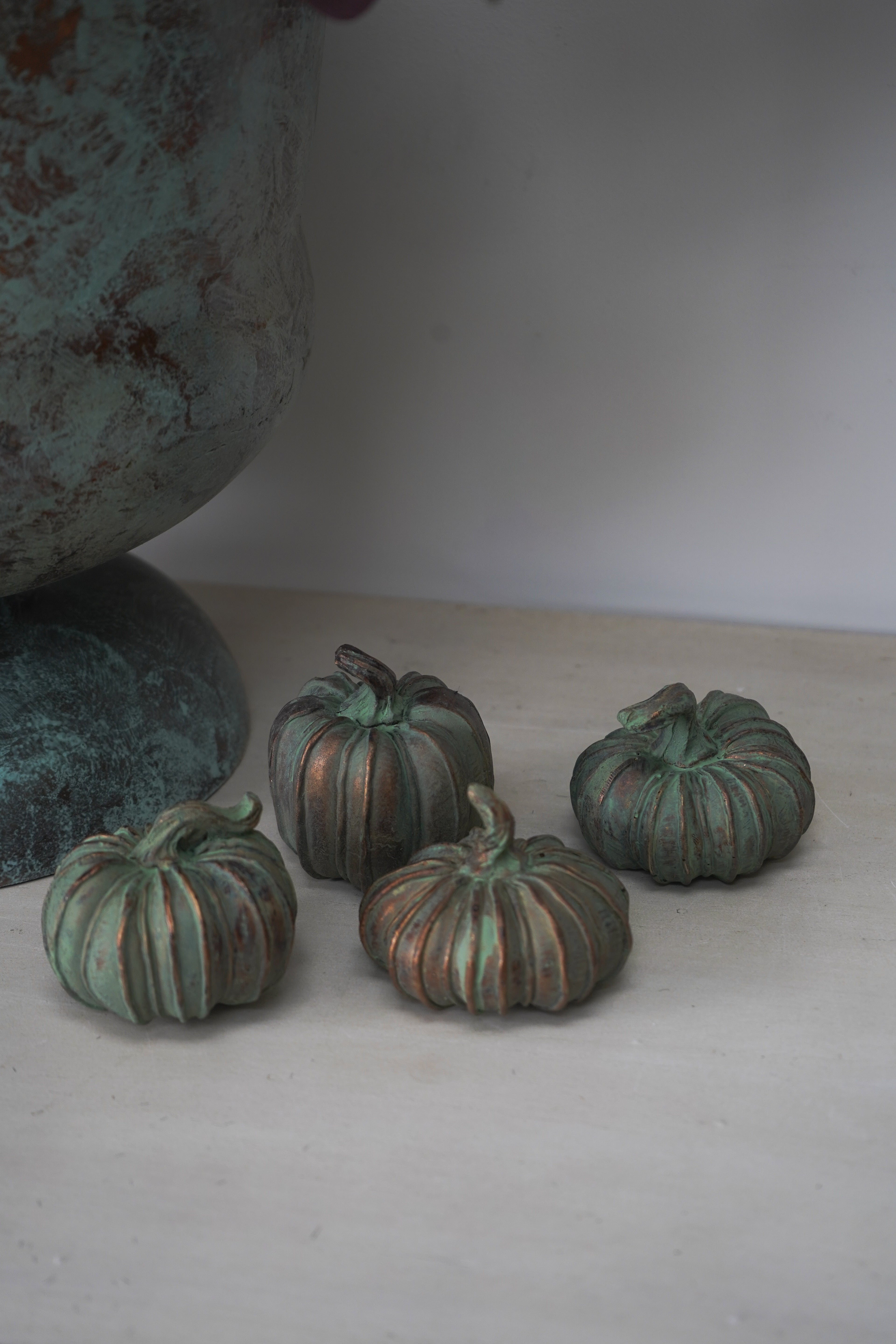 Four small green copper pumpkins on a white surface with a textured blue-green vase in the background.