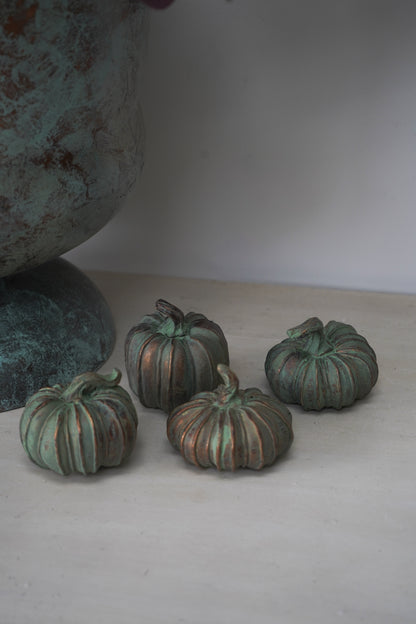 Four small green copper pumpkins on a white surface with a textured blue-green vase in the background.