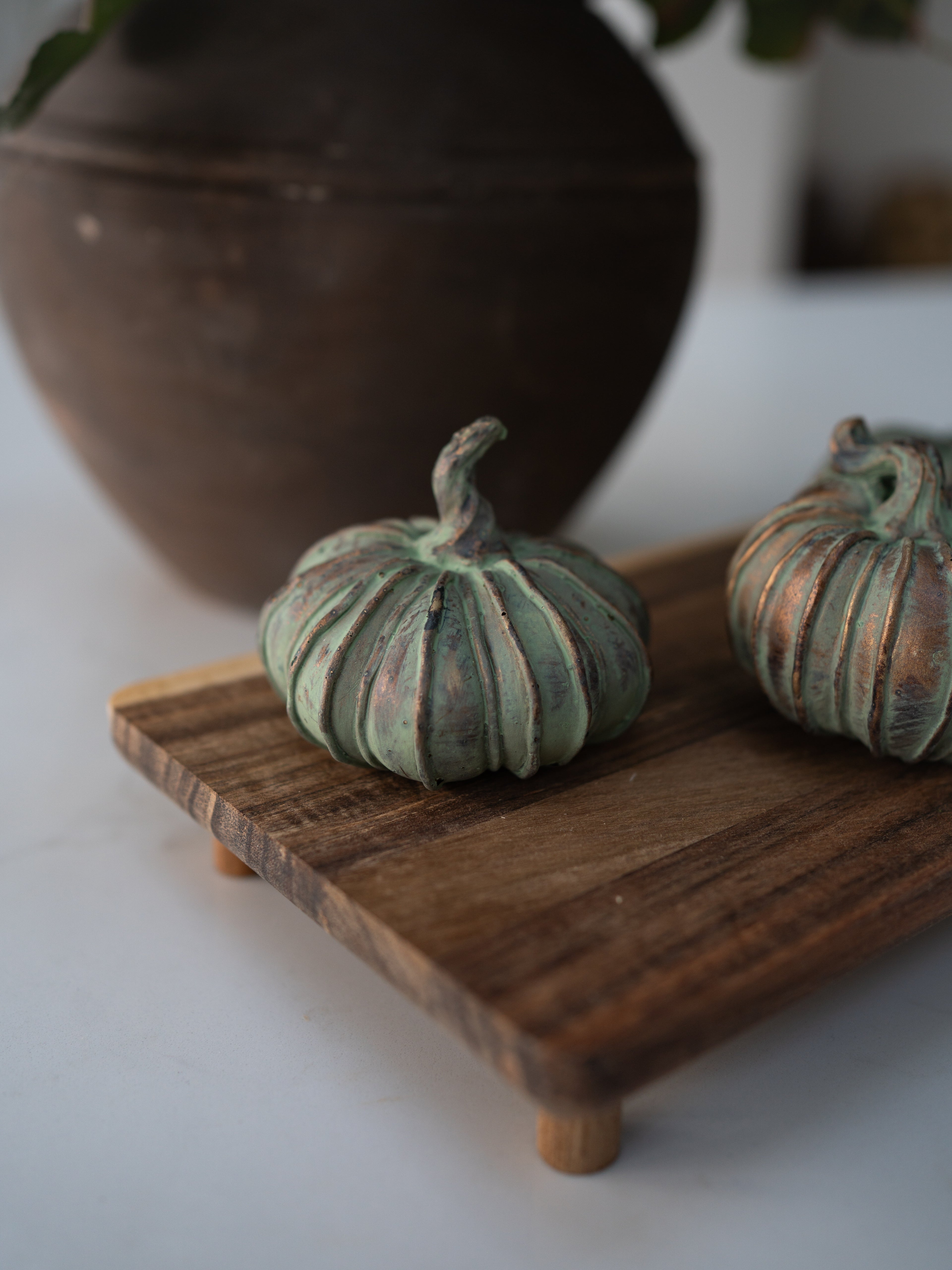 Two small decorative pumpkins with a distressed green and bronze patina finish, displayed on a wooden serving board with a blurred dark ceramic pot in the background.