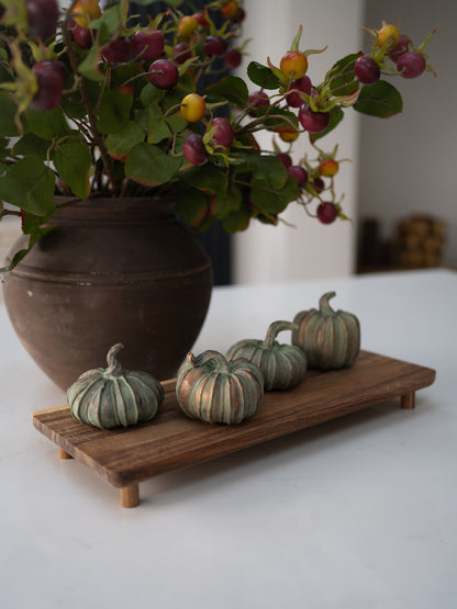 Four small decorative pumpkins with green and beige vertical stripes are arranged on a rectangular wooden tray, with a large brown ceramic vase of berries in the background.