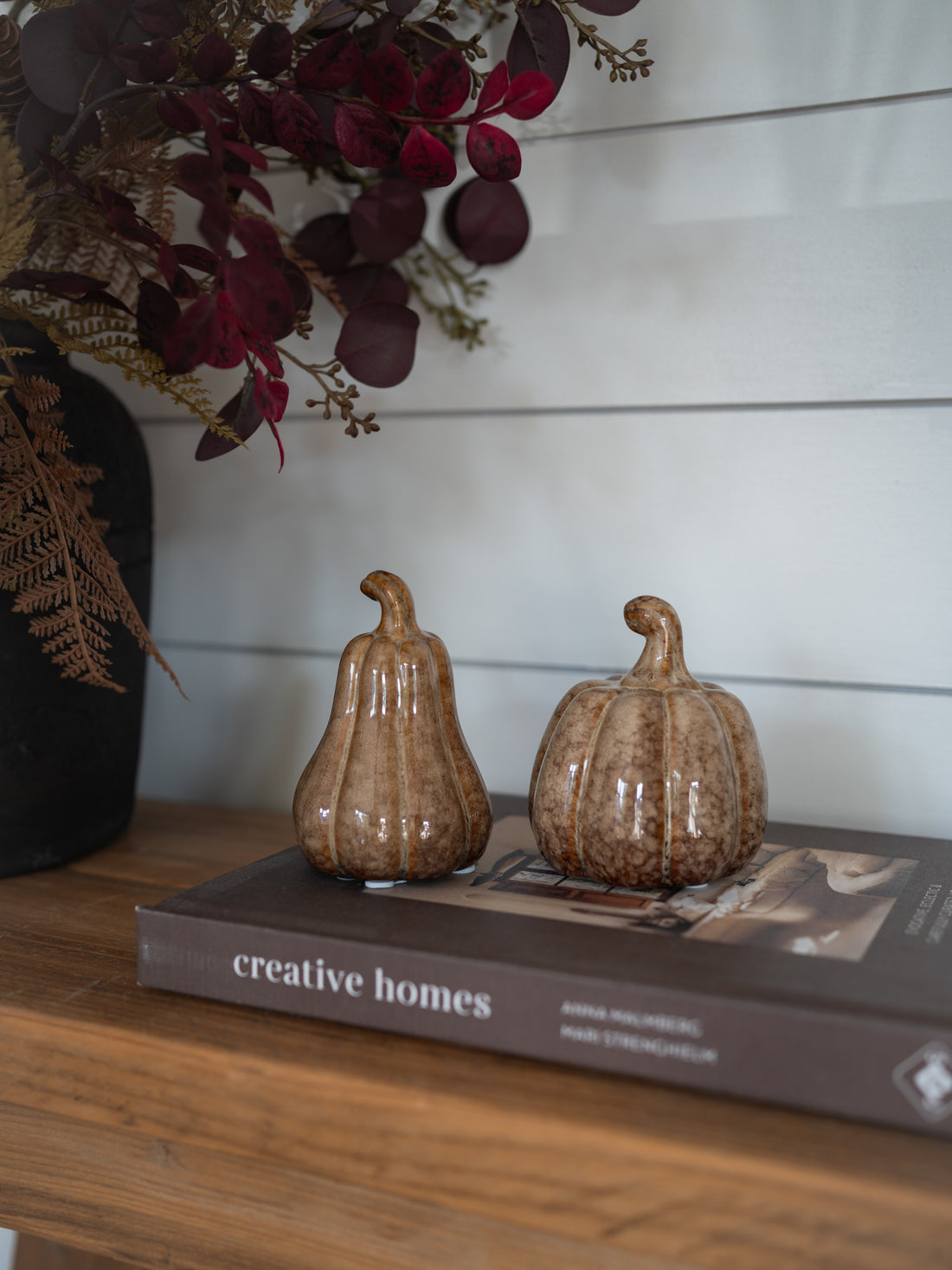 Two small ribbed ceramic pumpkins in a warm brown glaze sit on top of a book on a wooden shelf, with dried burgundy foliage and a white shiplap wall in the background.