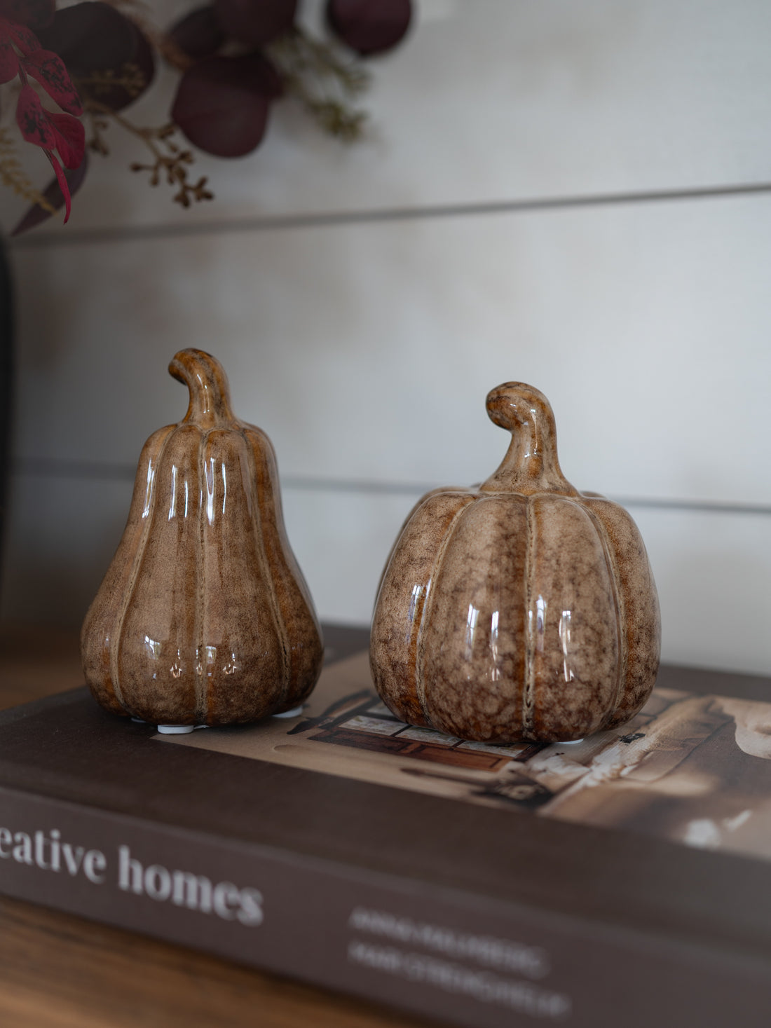 Two small glossy ceramic pumpkins in a mottled brown and beige glaze, displayed on top of a book in a home setting.