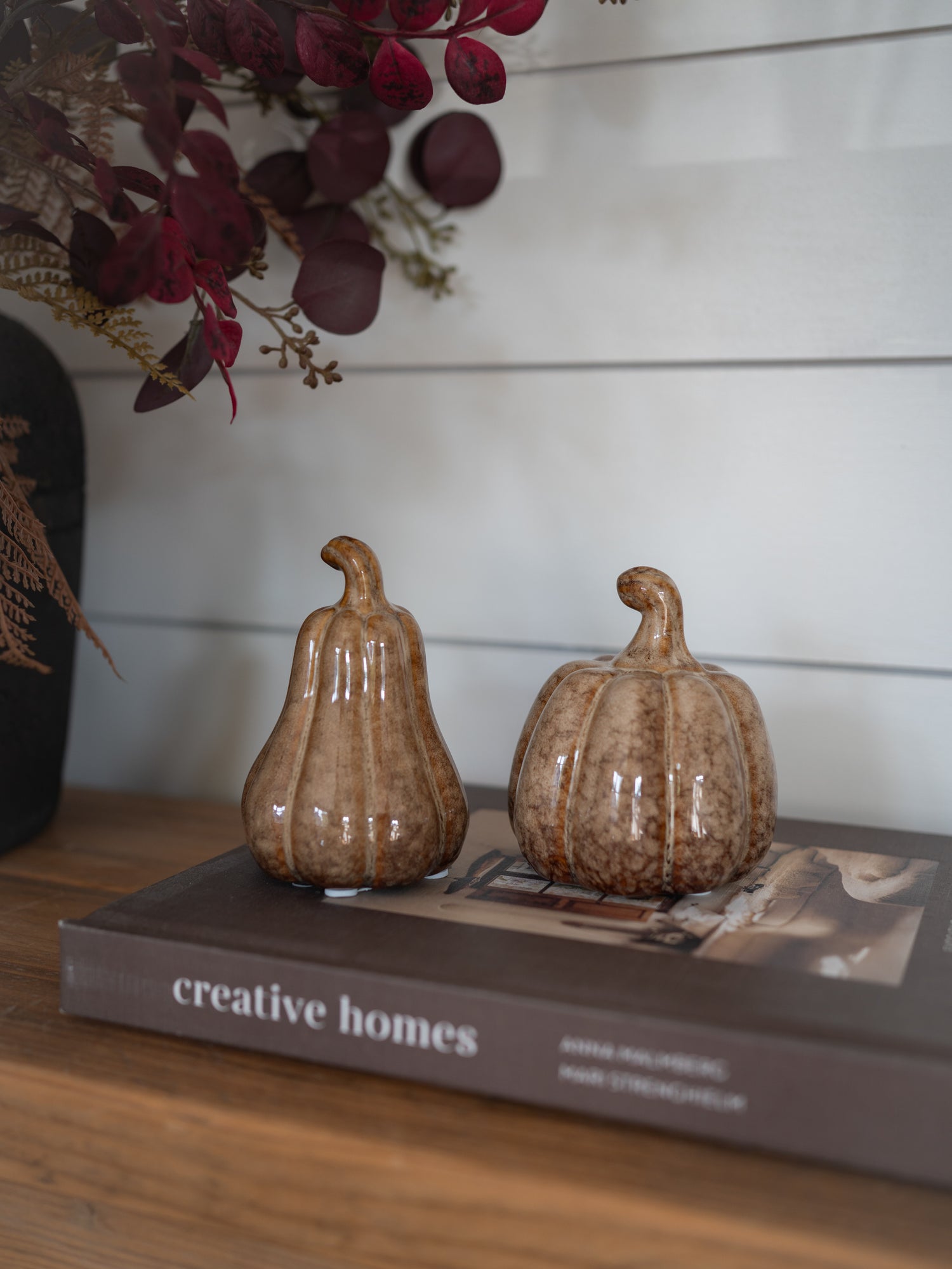 Two small decorative ceramic pumpkins in a brown speckled glaze, displayed on top of a book on a wooden surface against a white shiplap wall.