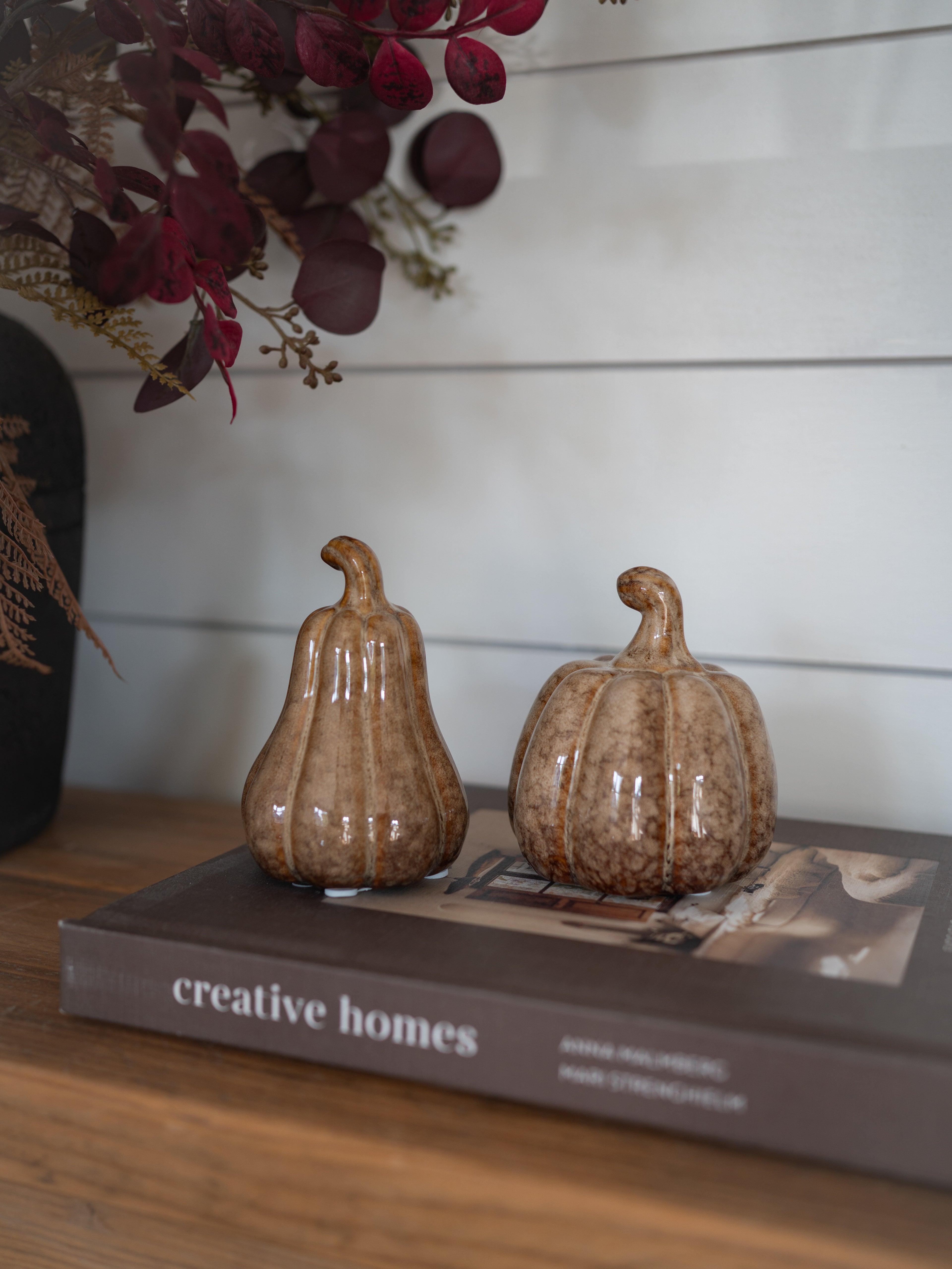 Two small decorative ceramic pumpkins in a brown speckled glaze, displayed on top of a book on a wooden surface against a white shiplap wall.