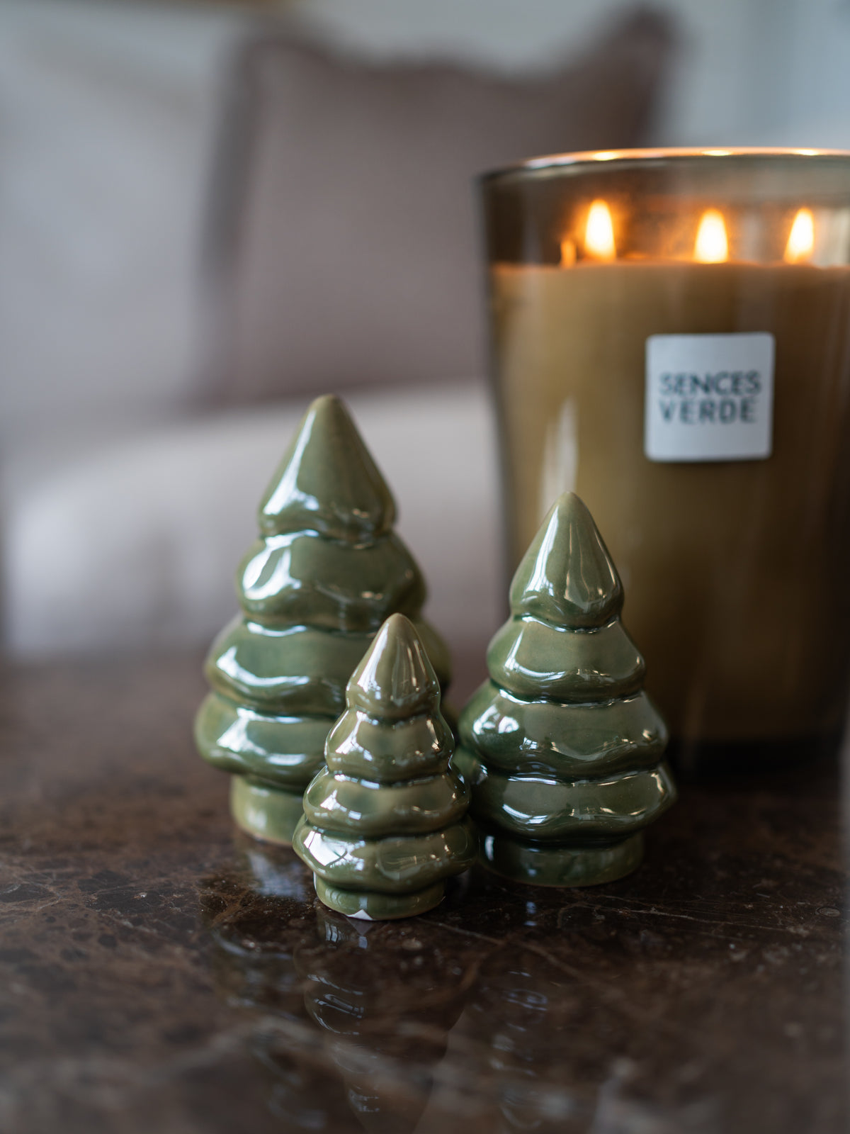 Three small glossy pearl-green ceramic Christmas tree ornaments standing on a dark marble surface, with a lit Sences Verde candle in a glass holder blurred in the background.