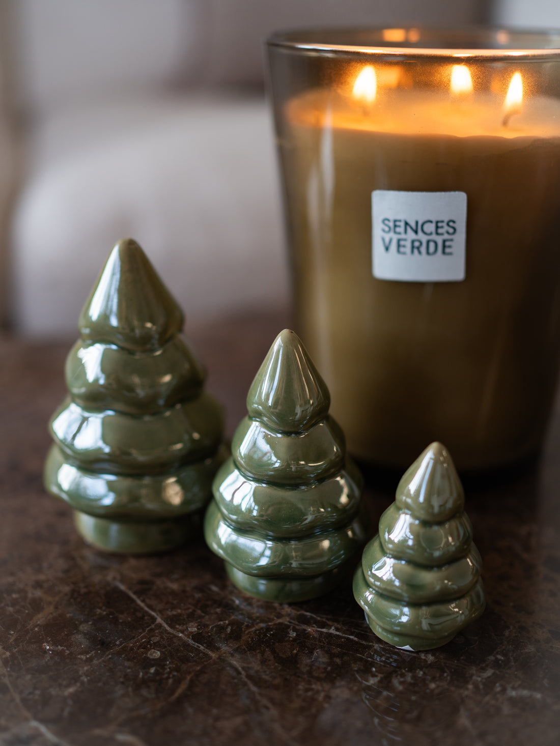 Three glossy olive green ceramic Christmas tree ornaments of varying sizes arranged on a dark marble surface in front of a lit candle in a glass jar. The candle label reads &