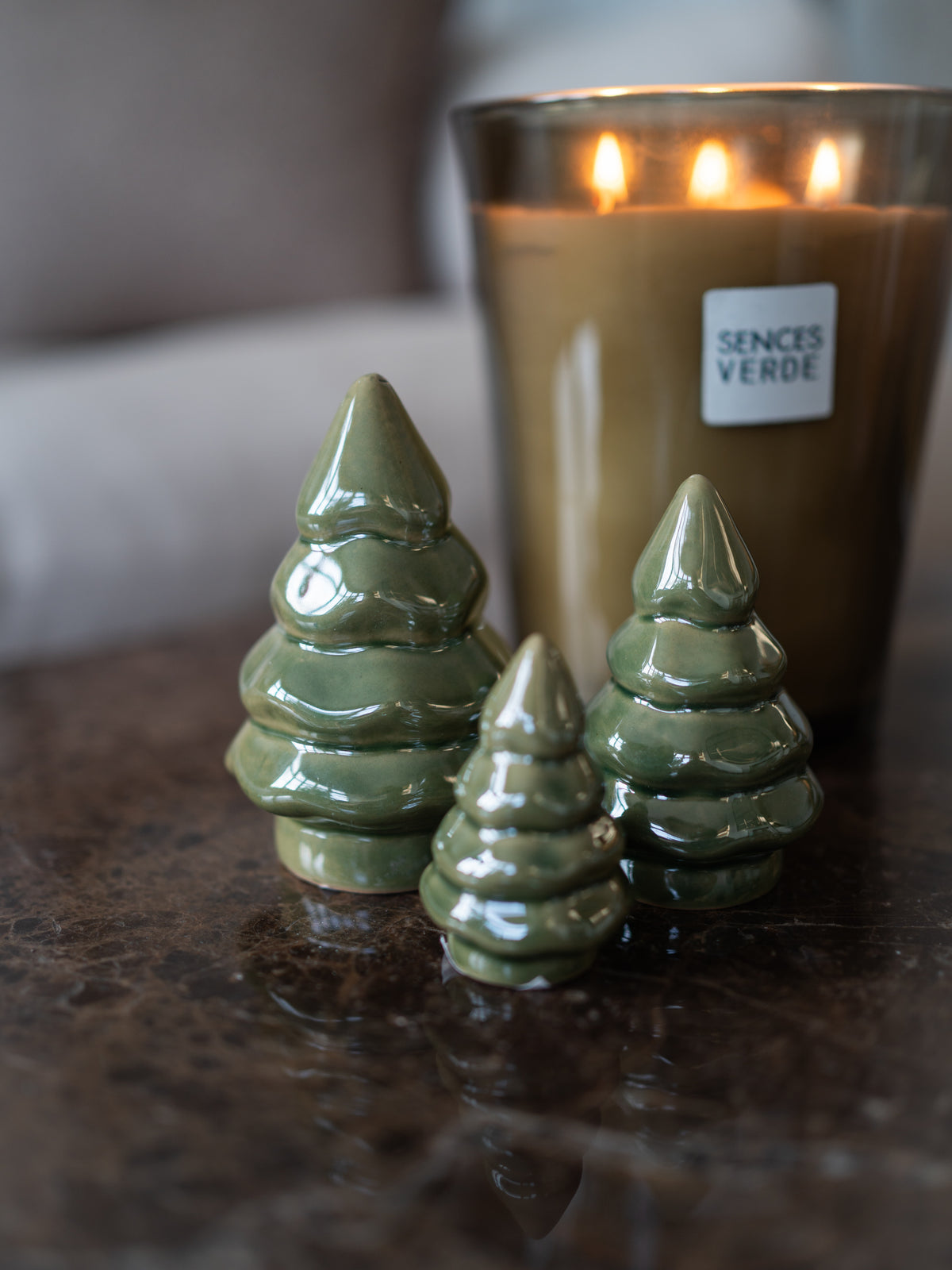 Three glossy olive green ceramic Christmas tree ornaments of varying sizes arranged on a dark reflective surface, with a blurred lit candle in the background. A label on the candle reads &