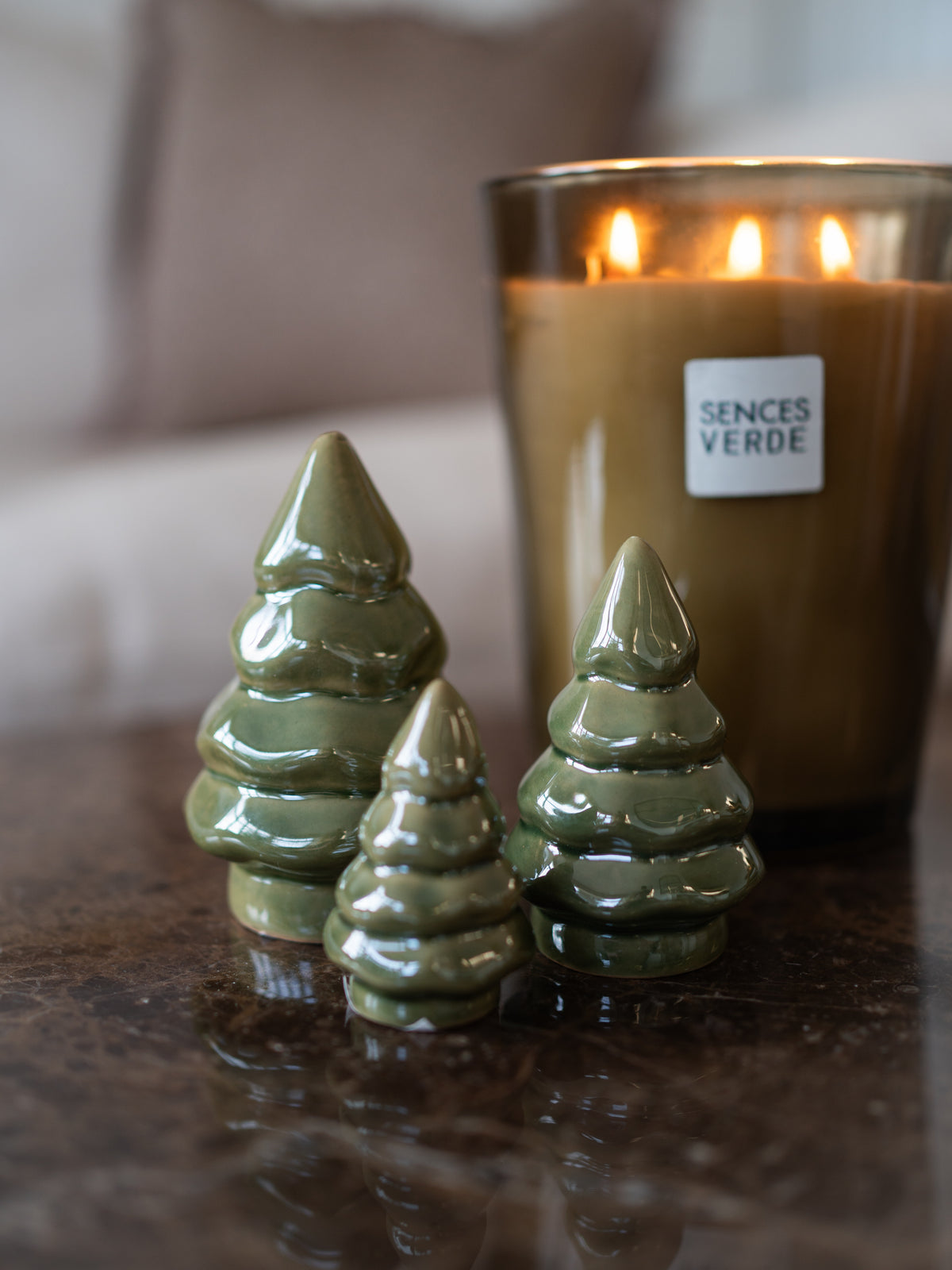 Three glossy pearl green ceramic Christmas tree ornaments on a dark reflective surface, with a lit Sences Verde candle in the blurred background.