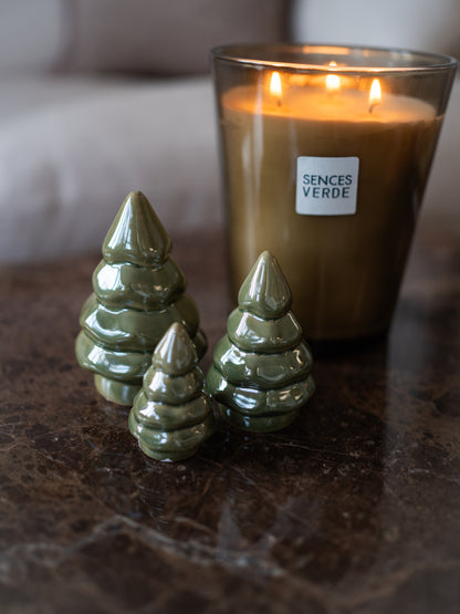 Three glossy pearl green ceramic Christmas tree ornaments arranged on a dark marble surface in front of a lit candle in a glass jar. The candle label reads &