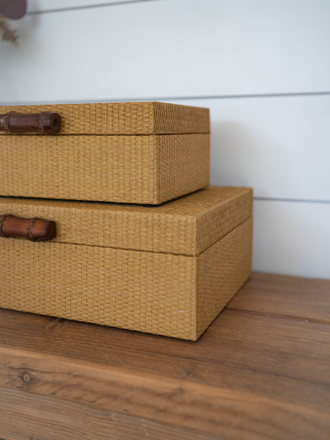 Two stacked woven storage boxes in a natural tan color with dark brown bamboo-style handles, resting on a wooden surface against a white paneled wall.