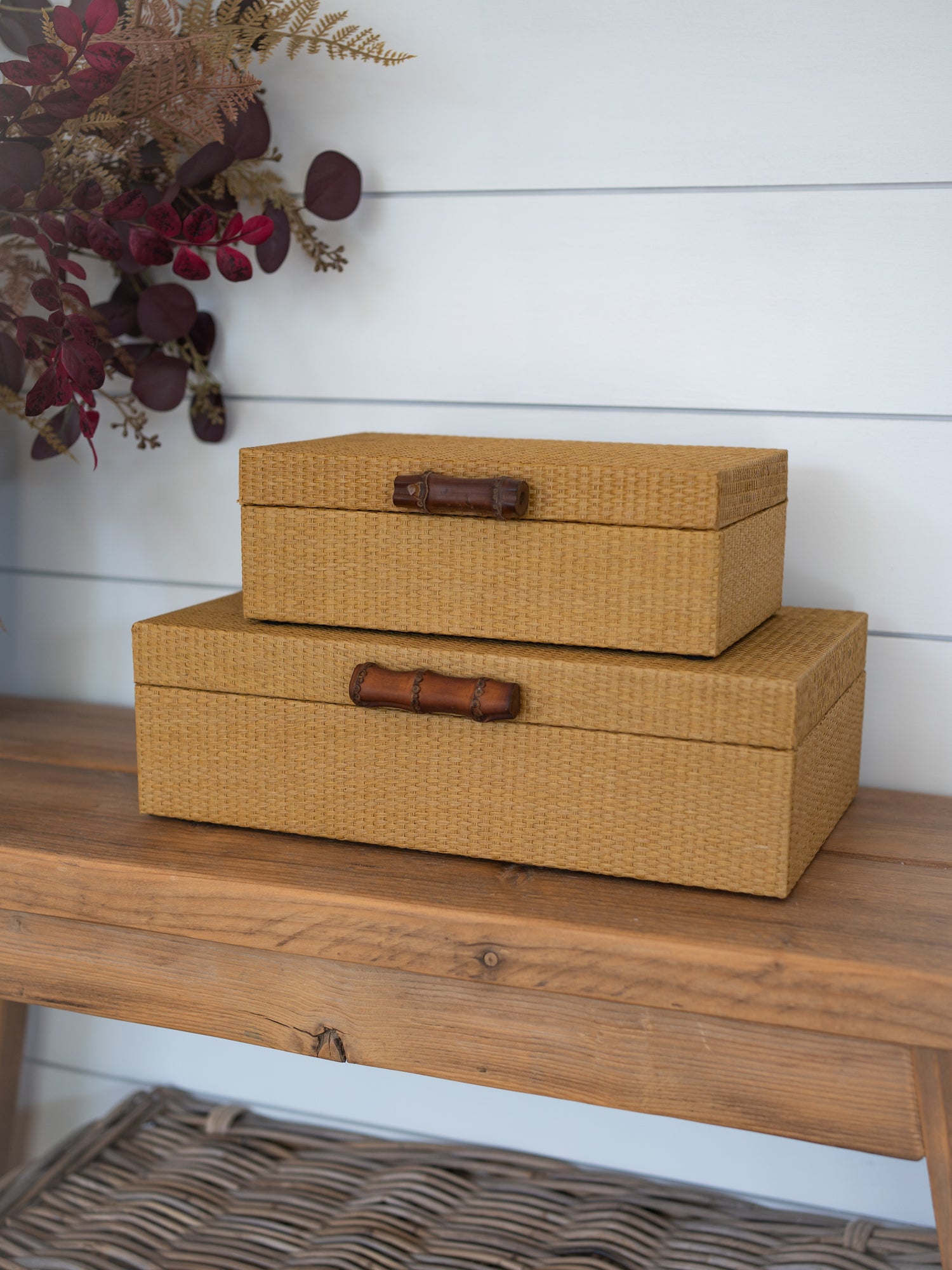 Two stacked woven storage boxes in a warm tan color with dark brown wooden bamboo handles, displayed on a rustic wooden bench against a white shiplap wall.