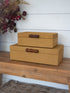 Two stacked woven storage boxes in a warm tan color with dark brown wooden bamboo handles, displayed on a rustic wooden bench against a white shiplap wall.