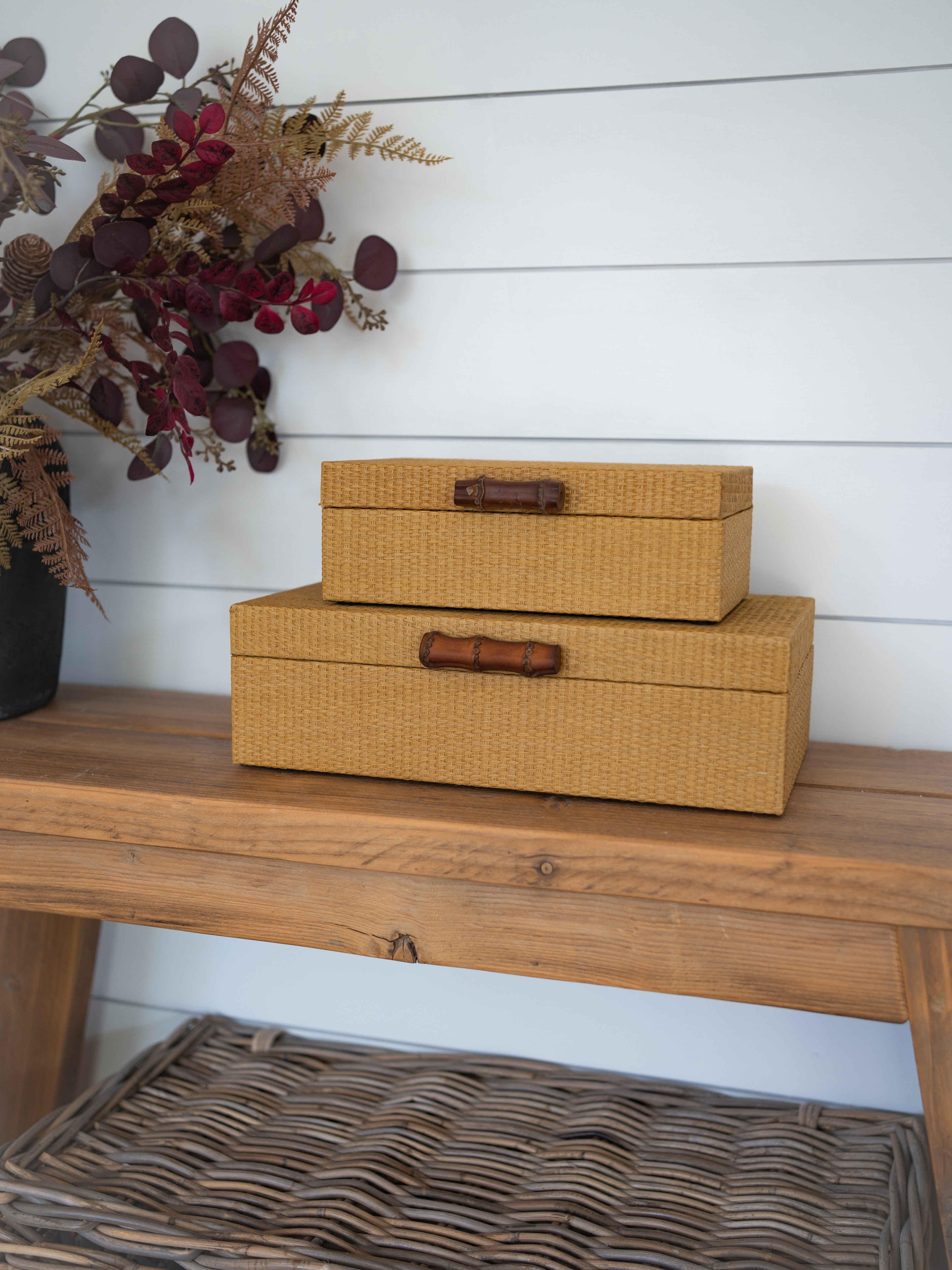 Two stacked rectangular storage boxes with woven mustard-yellow fabric covers and dark brown wooden handles, displayed on a wooden bench against a white shiplap wall.