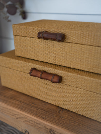 Two woven storage boxes stacked on a wooden surface, with a warm tan jute-like texture and dark brown bamboo-style handles.