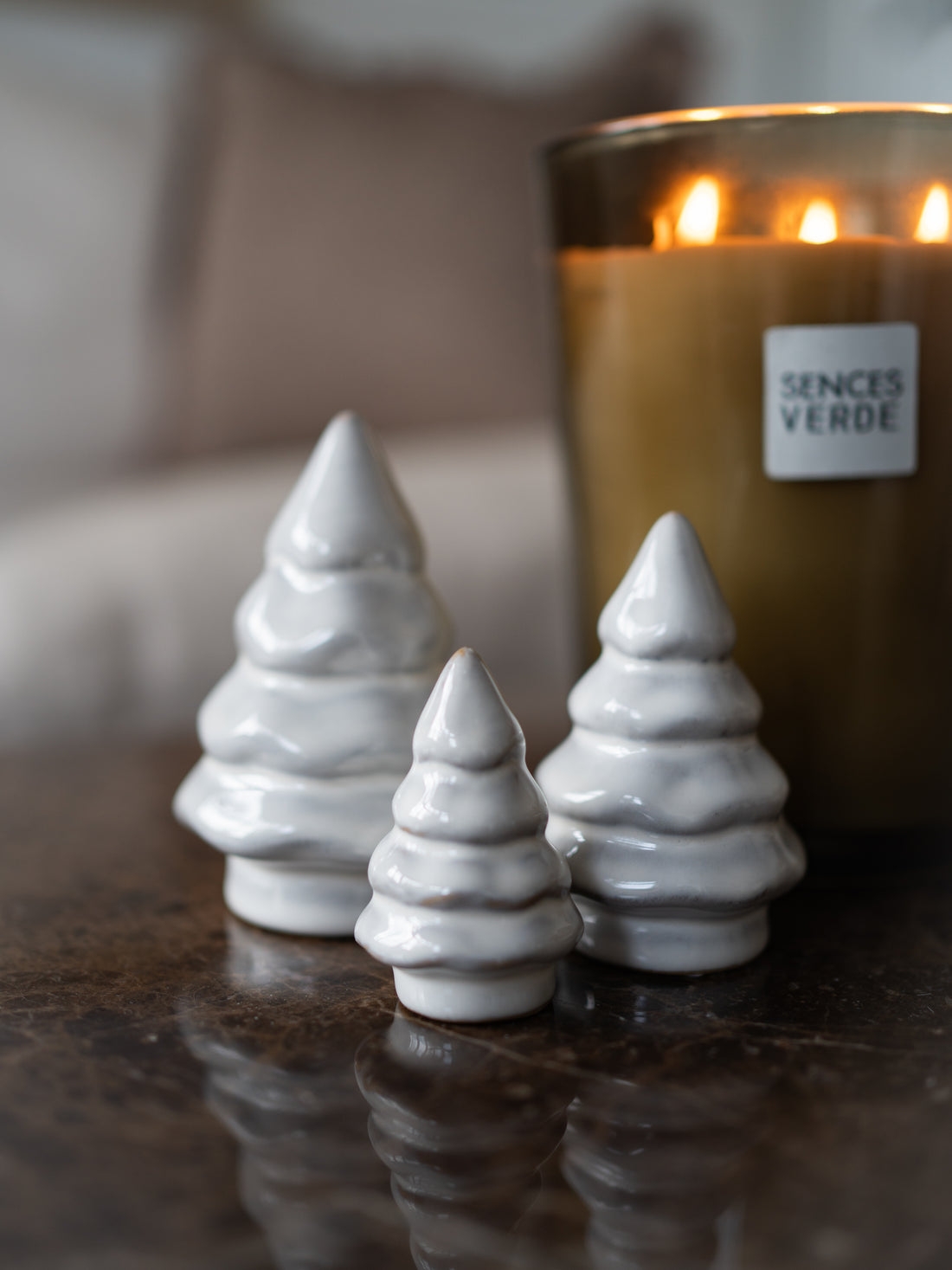Three small white ceramic Christmas tree ornaments with a glossy finish, displayed on a dark reflective surface next to a lit candle labeled &