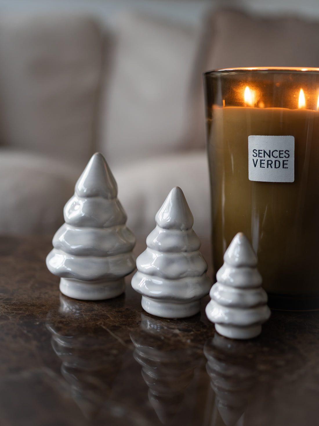 Three small white ceramic Christmas tree ornaments with a textured, layered design are arranged on a dark reflective surface next to a lit Senses Verde candle in a dark amber glass jar.