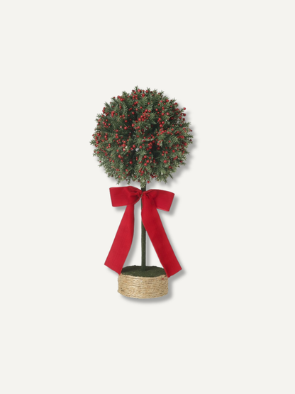 A festive artificial Christmas topiary tree with green foliage, red berries, and a large red ribbon bow, set in a woven basket pot against a light gray background.