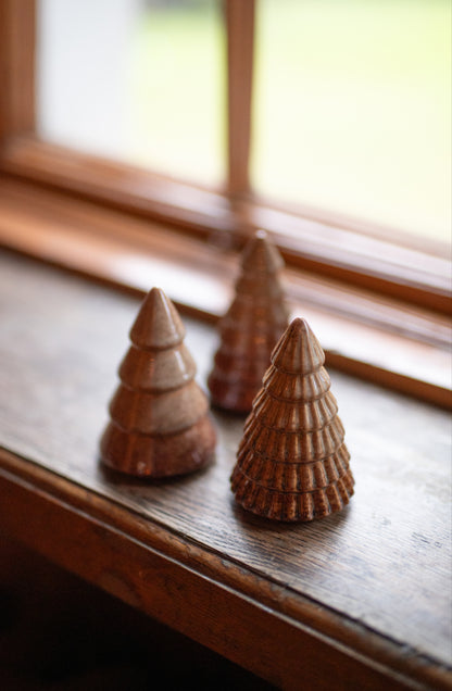Three small ceramic wooden Christmas tree ornaments in varying sizes, arranged on a dark wooden windowsill with a softly blurred window background.