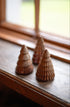 Three small ceramic wooden Christmas tree ornaments in varying sizes, arranged on a dark wooden windowsill with a softly blurred window background.
