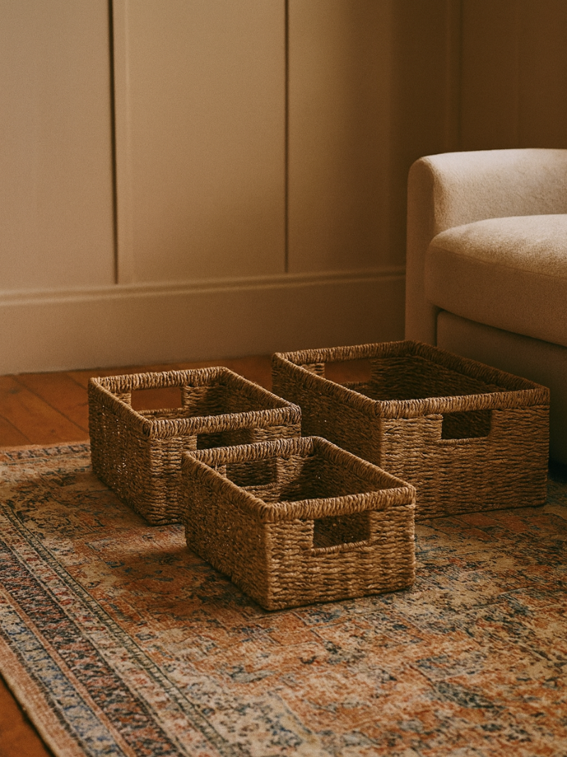 Three woven baskets on a patterned rug in a room with a beige wall and sofa.