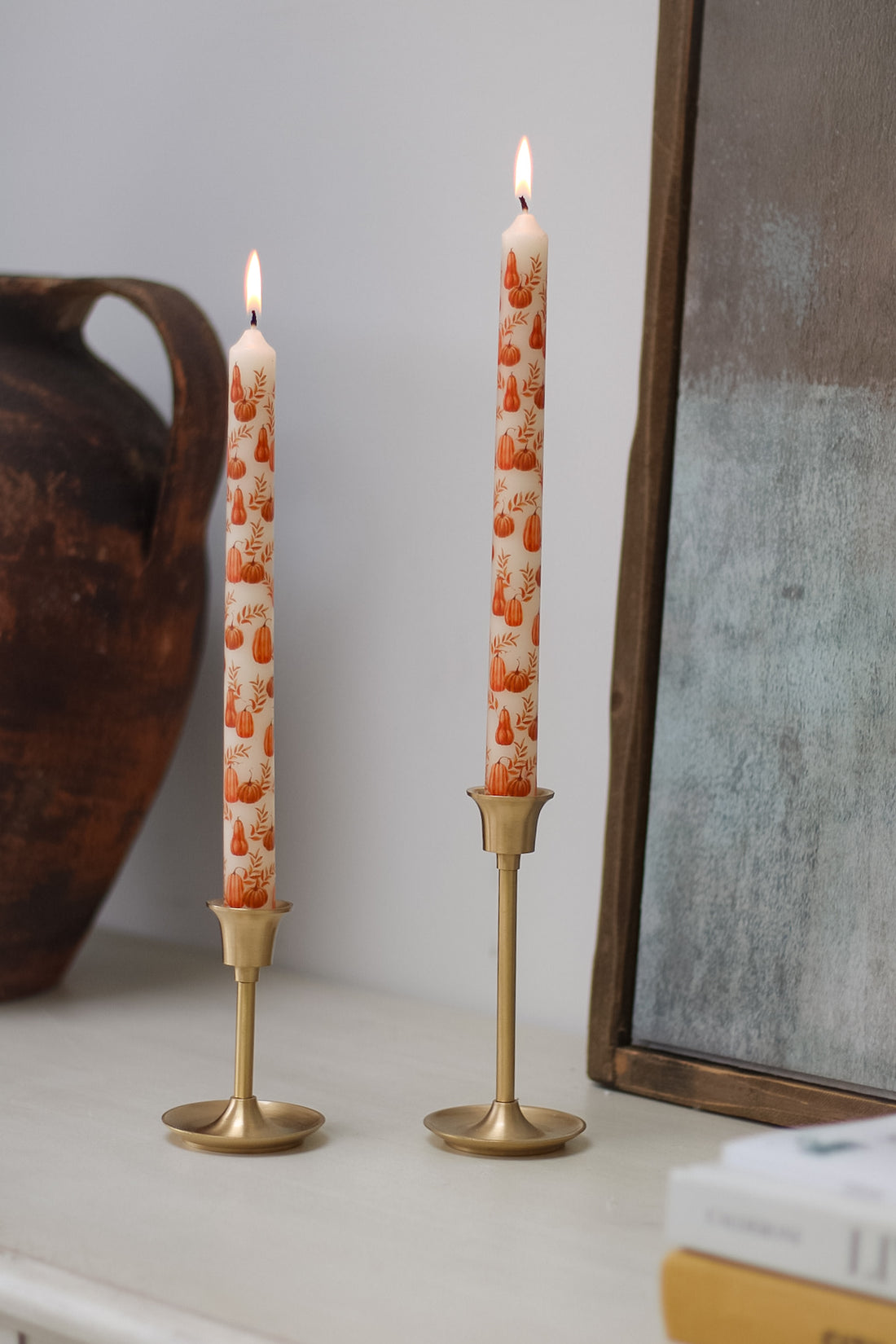 Two tall candles with pumpkin patterns in gold candle holders on a surface.