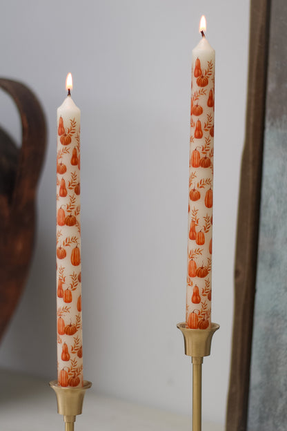 Two lit candles with pumpkin pattern in gold holders against a neutral background