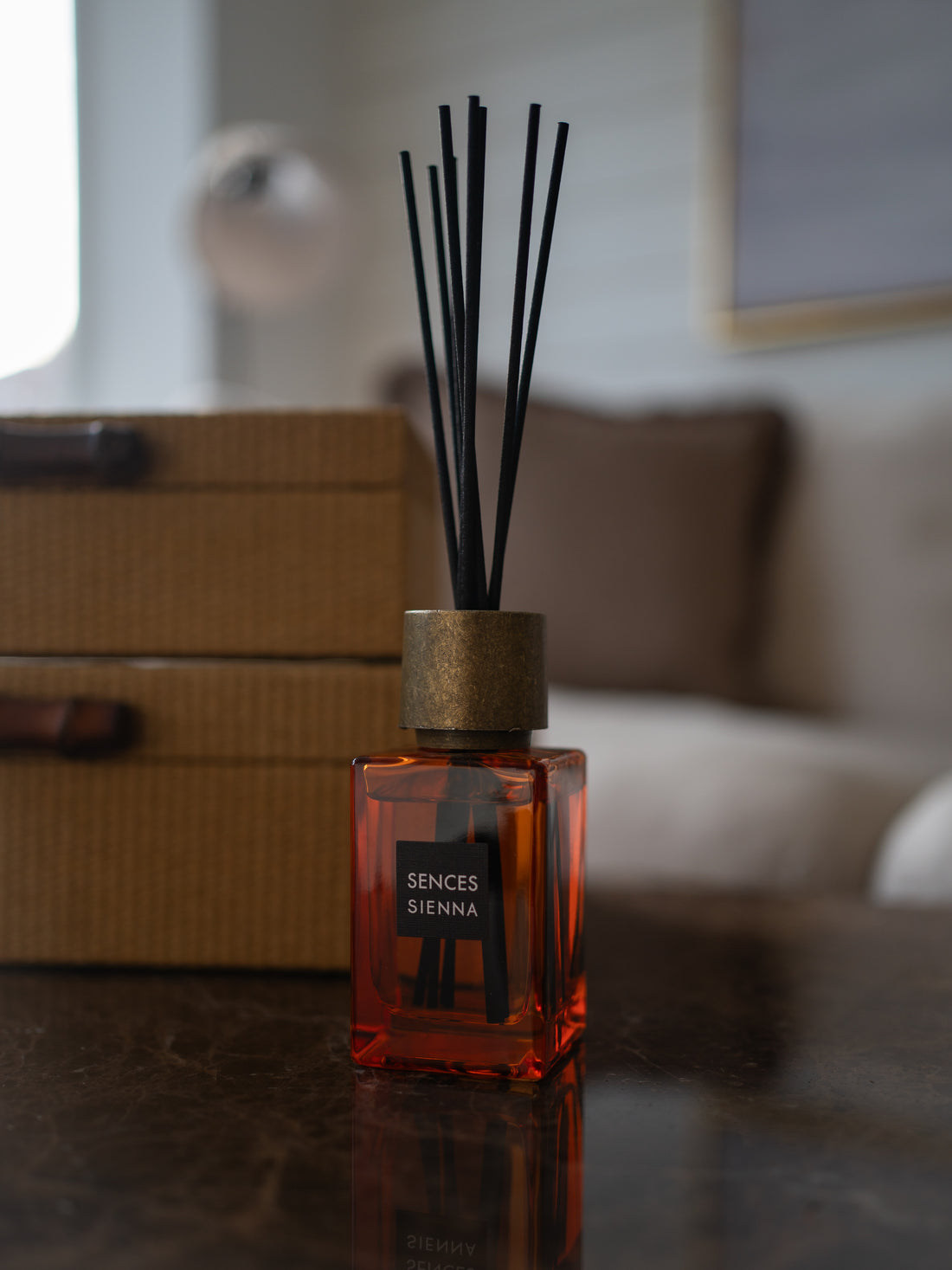 A Sences Sienna reed diffuser bottle made of transparent amber glass with black reeds, set on a dark reflective surface with a blurred home interior background.