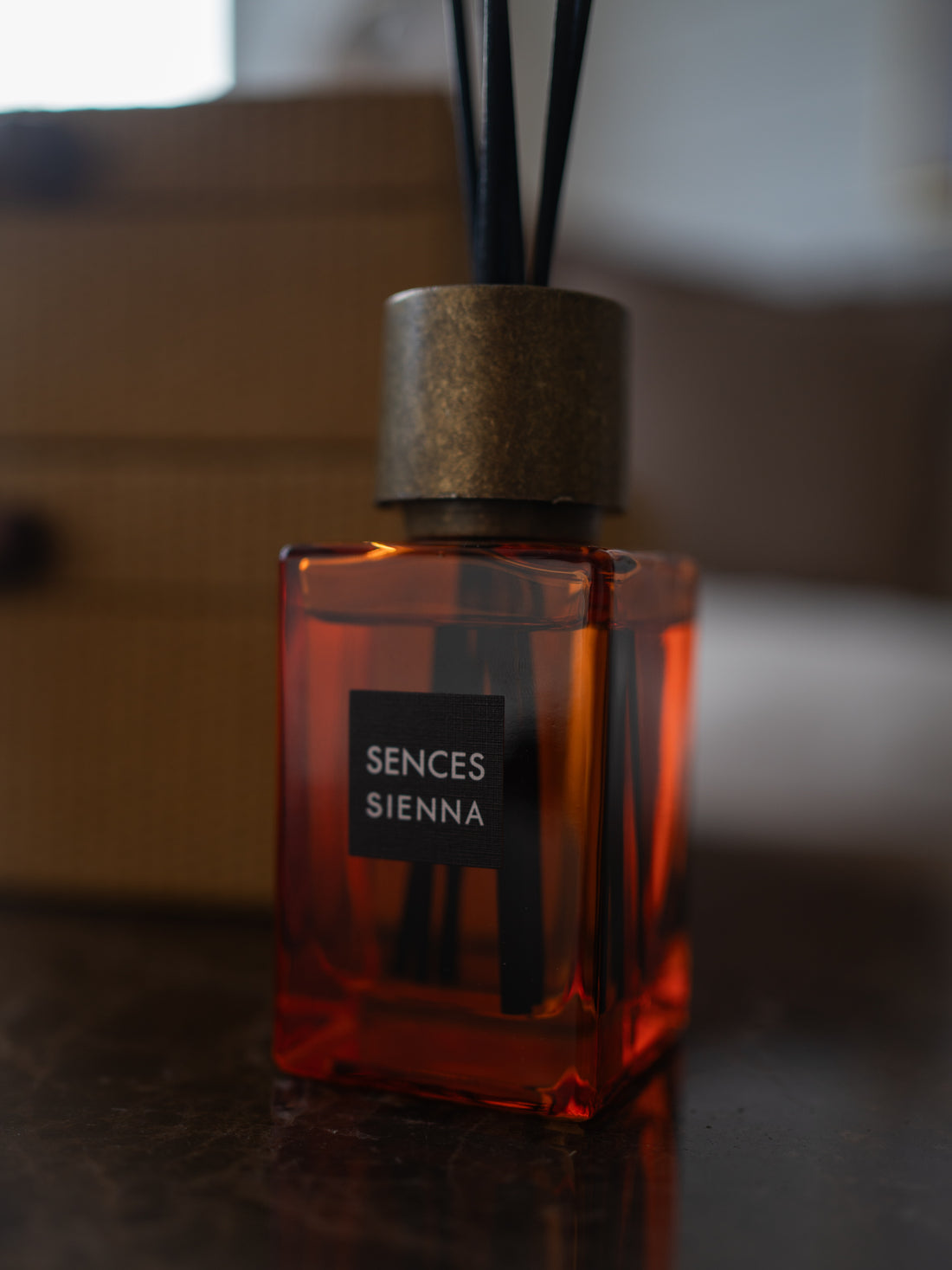 A Sences Sienna reed diffuser bottle with amber-colored liquid, a textured metallic cap, and black reeds, set on a dark reflective surface with a blurred background.