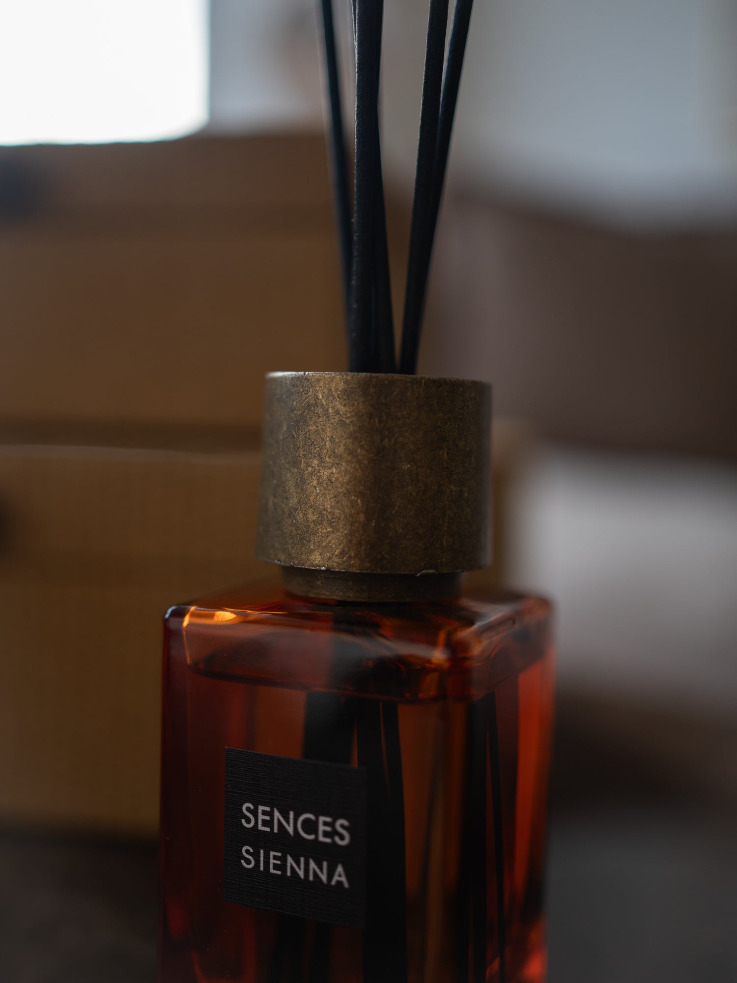 A close-up of a Sences Sienna reed diffuser bottle with amber-colored liquid, a textured metallic gold cap, and black reeds, set against a softly blurred background.