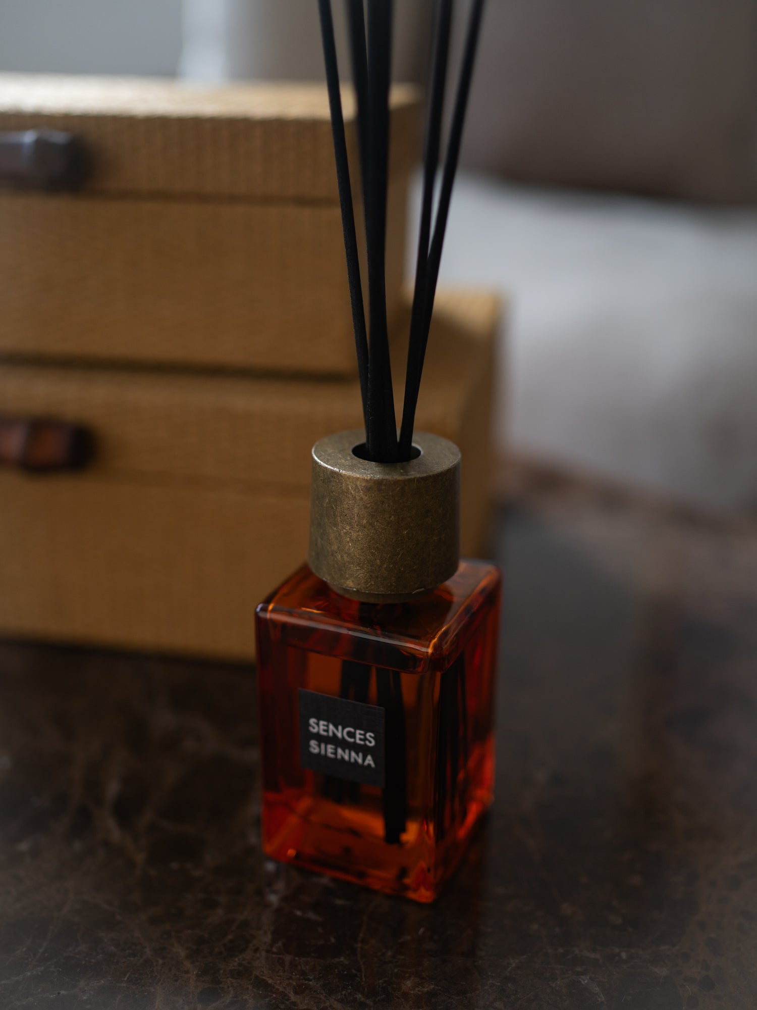 A reed diffuser bottle with amber-colored liquid, a brass cap, and black reeds, labeled &