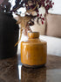 A small glazed ceramic bottle with a mustard yellow finish and a dark brown base, styled on a polished dark marble tabletop next to a black vase with dried foliage.