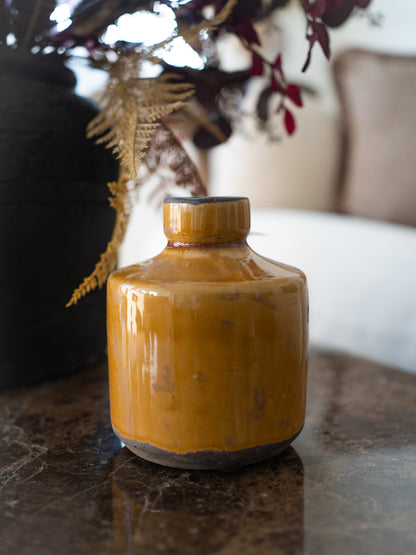A small glazed ceramic vase with a mustard yellow finish and a dark base, styled on a dark marble tabletop with dried ferns in the background.