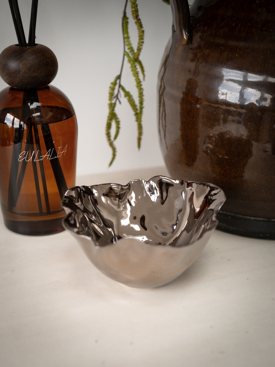 Decorative items including a metallic bowl, luxury reel diffuser, and a brown ceramic vase on a light background.
