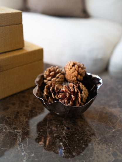 A small dark copper metal bowl filled with natural pinecones, placed on a polished dark marble surface with stacked tan boxes and a softly blurred sofa in the background.