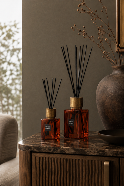 Two amber glass reed diffusers with black reeds and gold caps sit on a dark marble-topped cabinet in a styled interior setting.