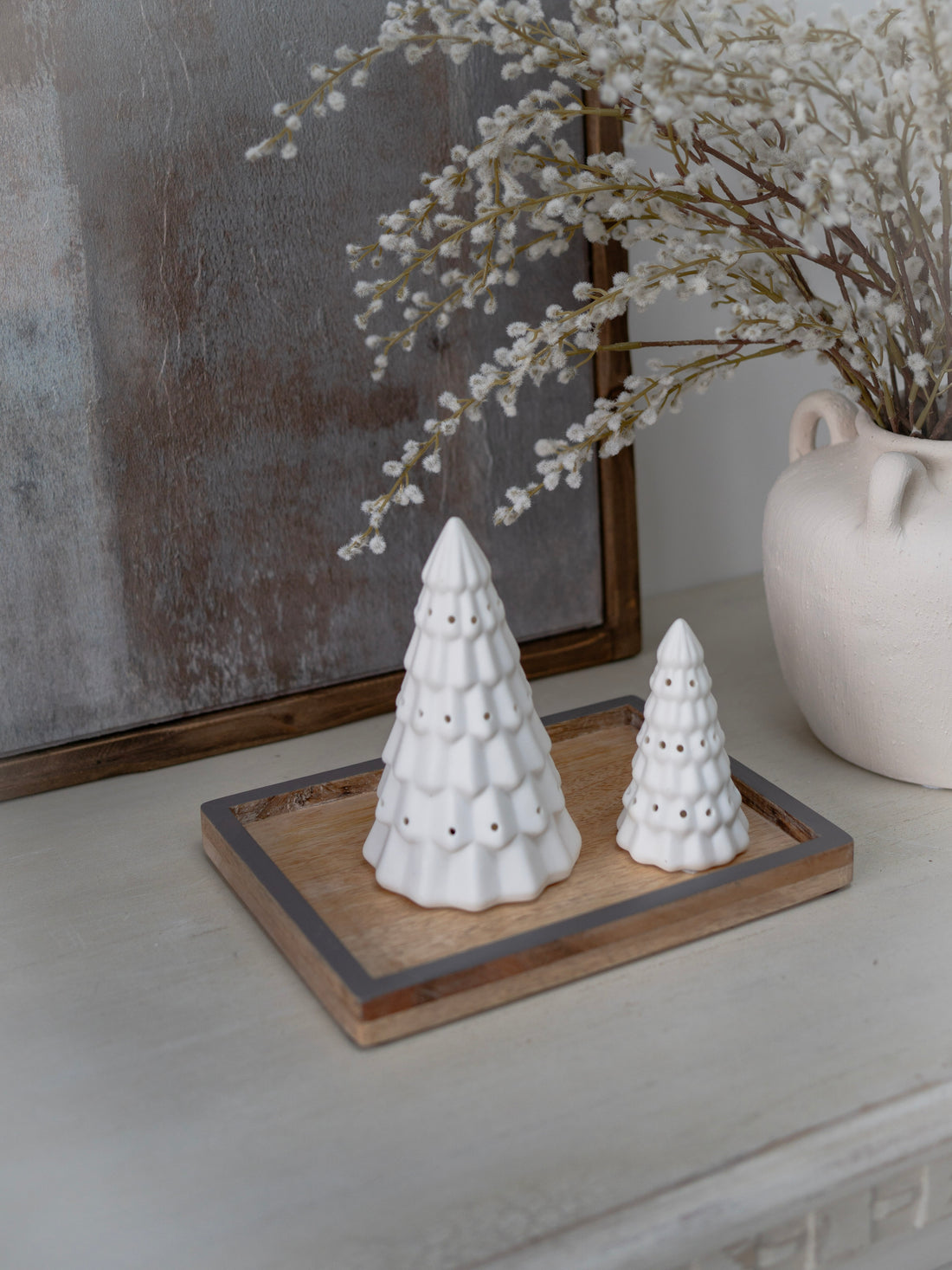 Two white ceramic trees on a wooden tray with a vase and branches in the background.
