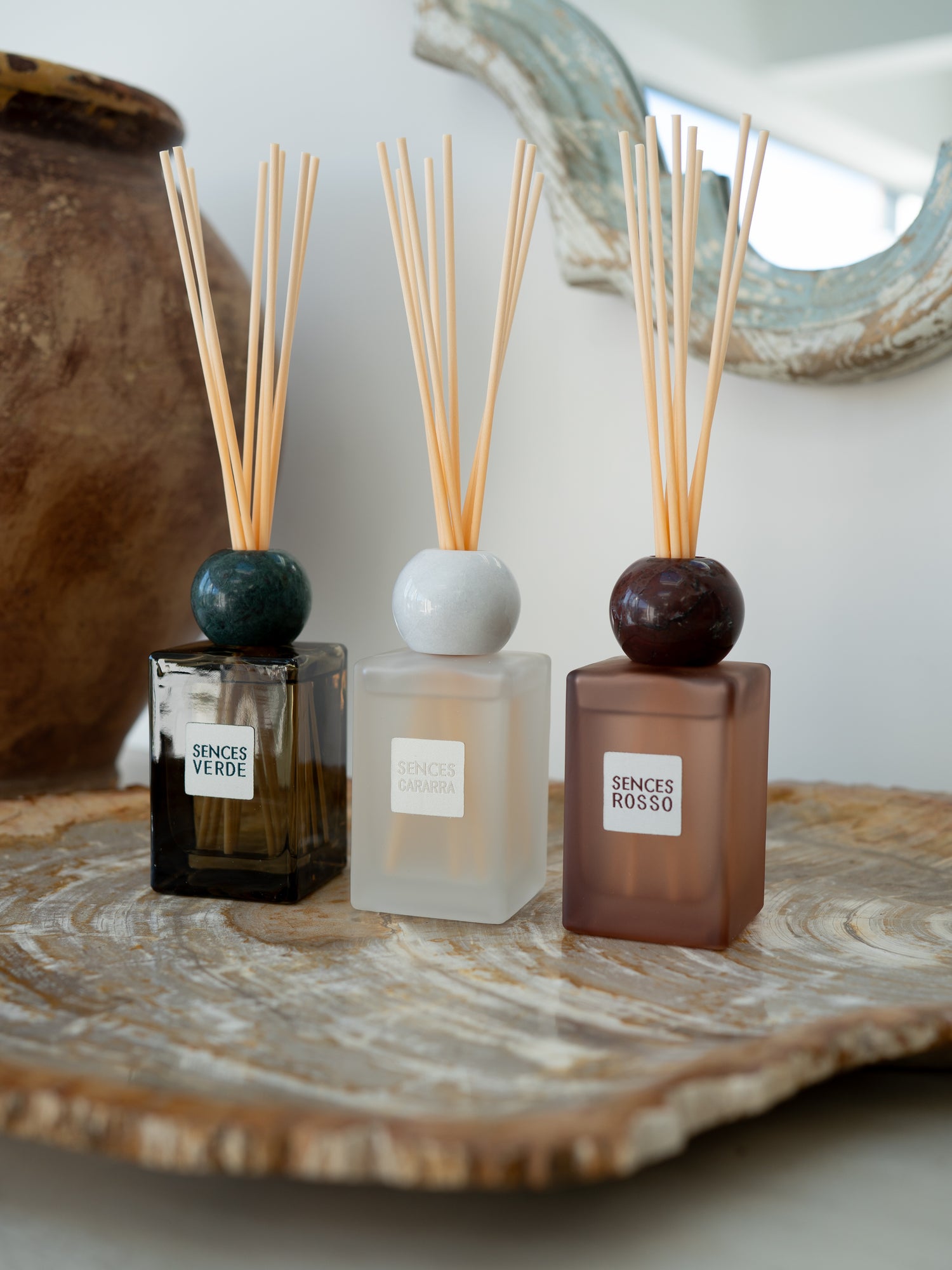 Three reed diffusers with wooden reeds are arranged on a rustic wood slice tray. The diffusers have square glass bottles in black, frosted white, and smoky brown, topped with round dark green, white, and dark reddish-brown stoppers.