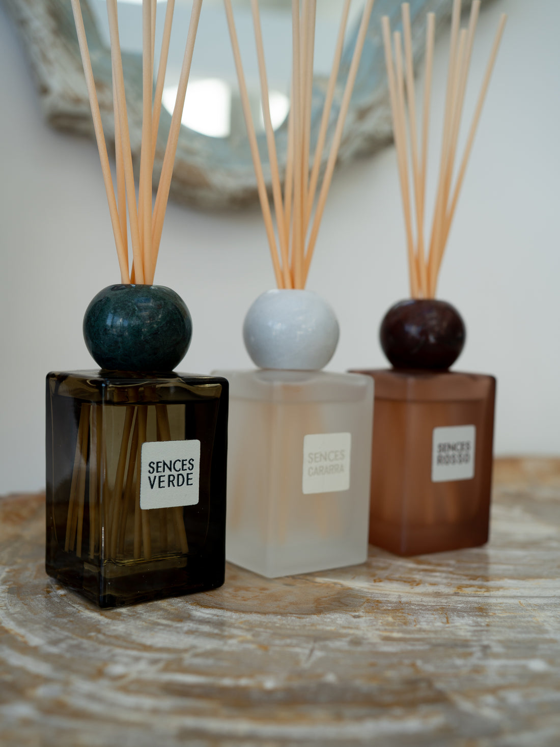 Three reed diffusers with square glass bottles and natural reed sticks are arranged on a stone surface. The left bottle is dark amber with a dark green round top and a white label reading &