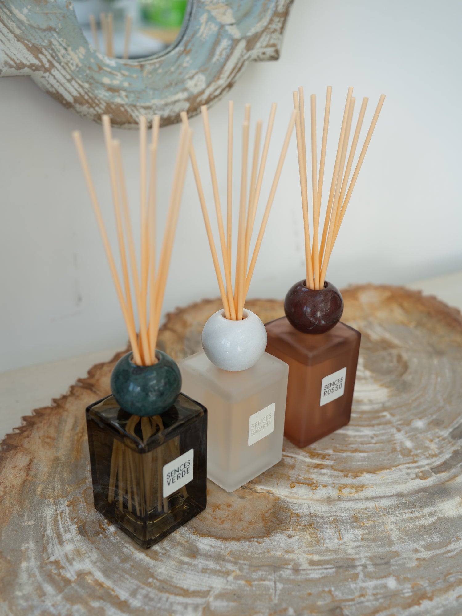 Three reed diffusers with wooden reeds in square glass bottles of black, frosted white, and terracotta brown, arranged on a round wood slice tray.