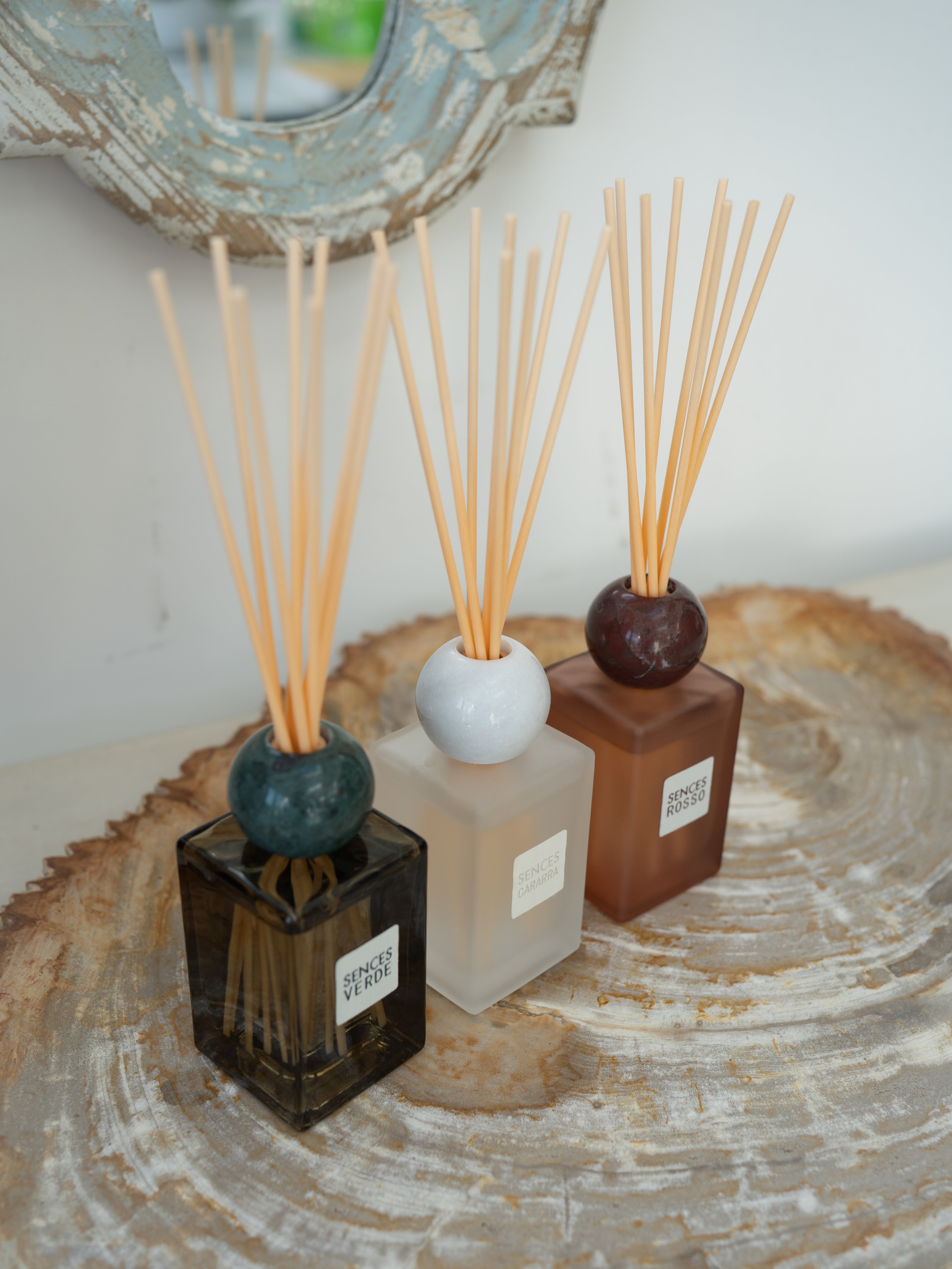 Three reed diffusers with wooden reeds in square glass bottles of black, frosted white, and terracotta brown, arranged on a round wood slice tray.
