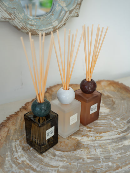 Three reed diffusers with wooden reeds in square glass bottles of black, frosted white, and terracotta brown, arranged on a round wood slice tray.