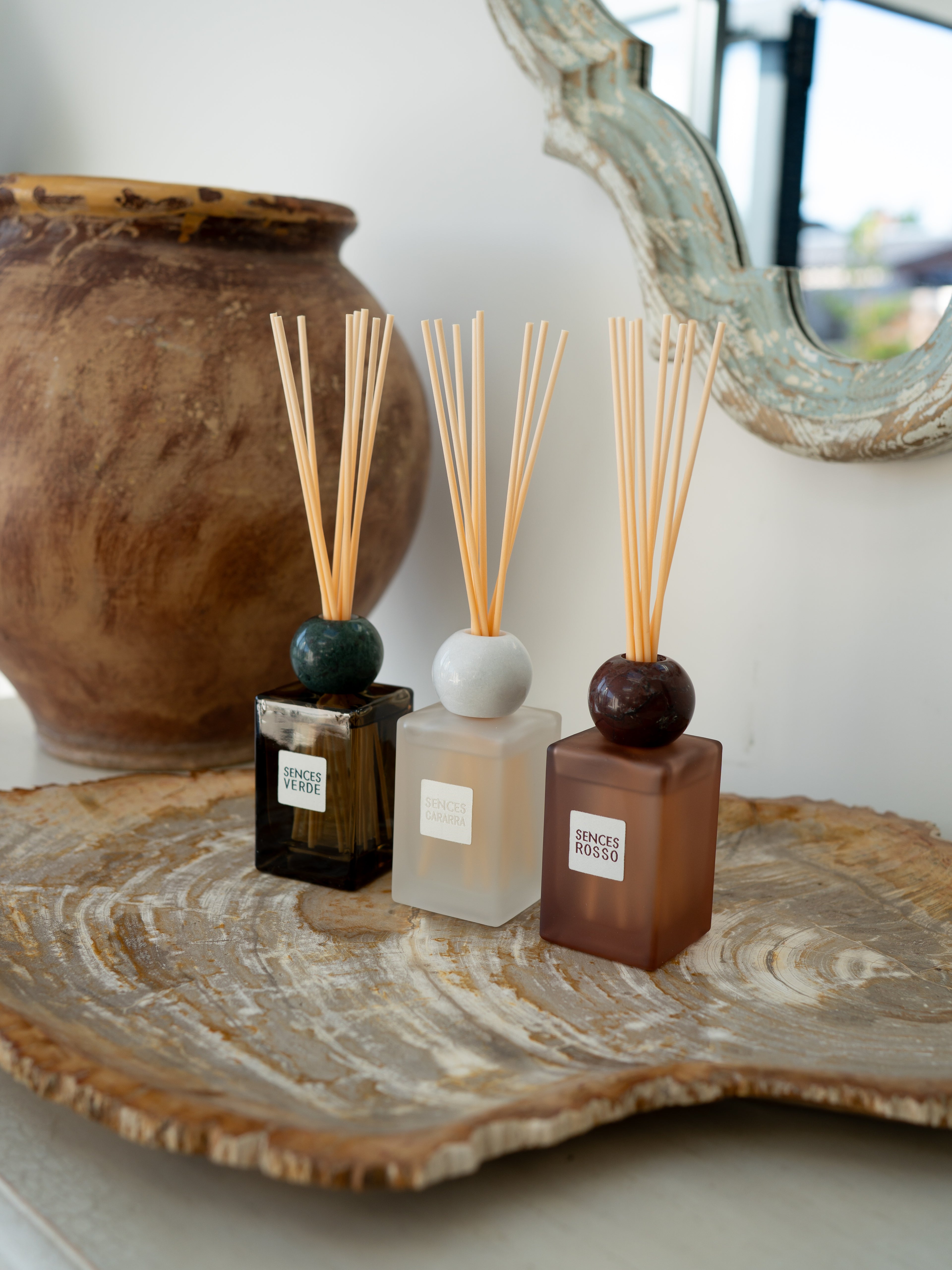 Three reed diffusers with wooden reeds are arranged on a rustic wood slice tray. The diffusers have frosted glass bottles in dark amber, translucent white, and dark brown, topped with dark green, white, and dark brown spherical caps.