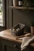 A woven wicker basket sits on a wooden countertop in a styled kitchen or bathroom setting, alongside a wooden brush and a textured beige towel. The background features a dark gray wall, a wooden shelf with a vase and plant, and a window on the left.