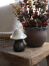 A small ceramic mushroom-shaped decor piece with a glossy white cap and dark glazed base, displayed on a rustic wooden table next to a potted plant.