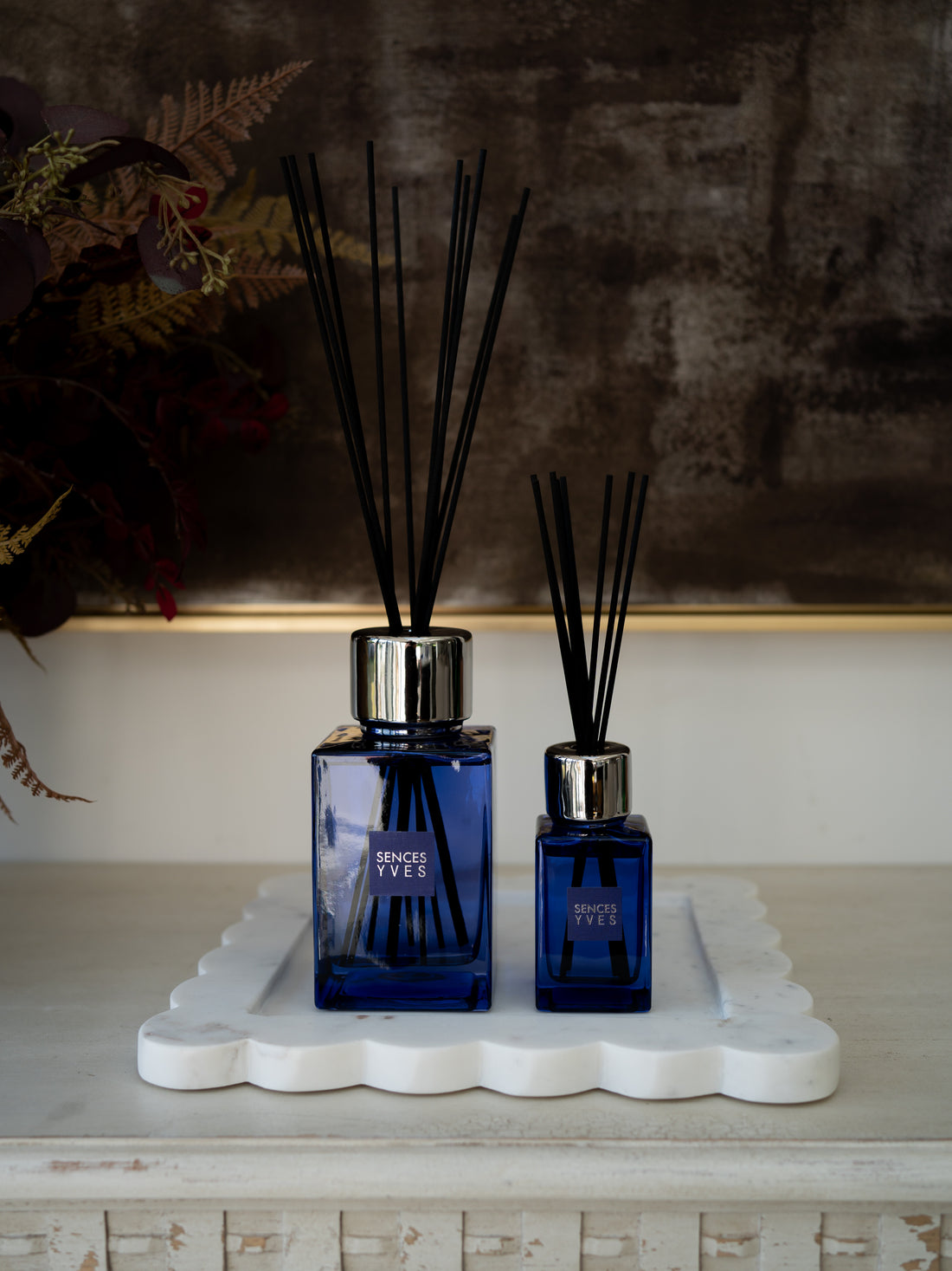 Two cobalt blue glass reed diffusers with black reeds and silver caps, displayed on a white scalloped tray on a rustic mantelpiece.