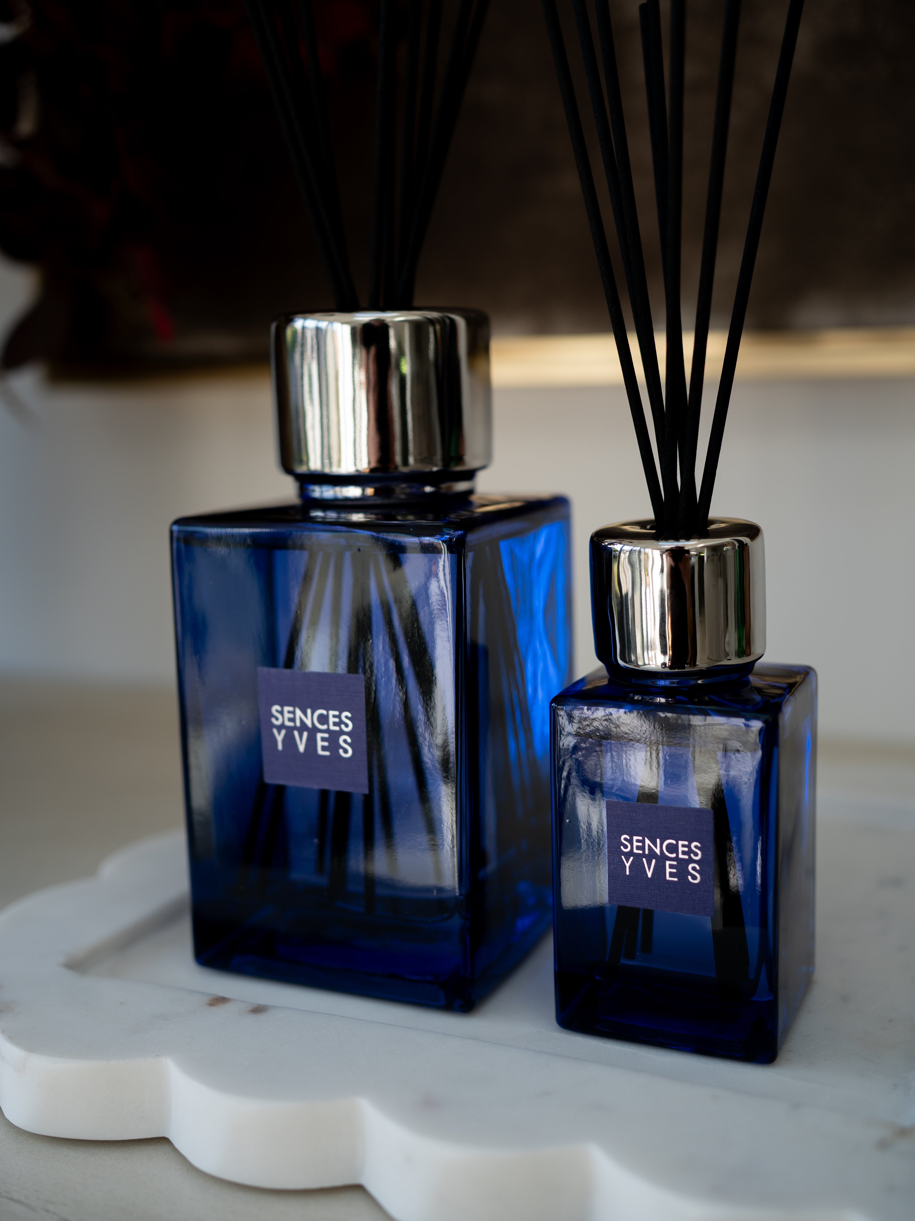 Two Scentes Yves reed diffusers in deep cobalt blue glass bottles with silver caps and black reeds, displayed on a white scalloped tray.