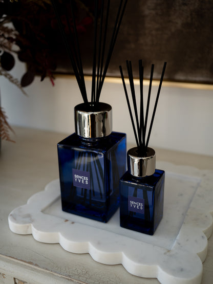 Two blue glass reed diffusers with black reeds and silver caps, displayed on a white scalloped tray.
